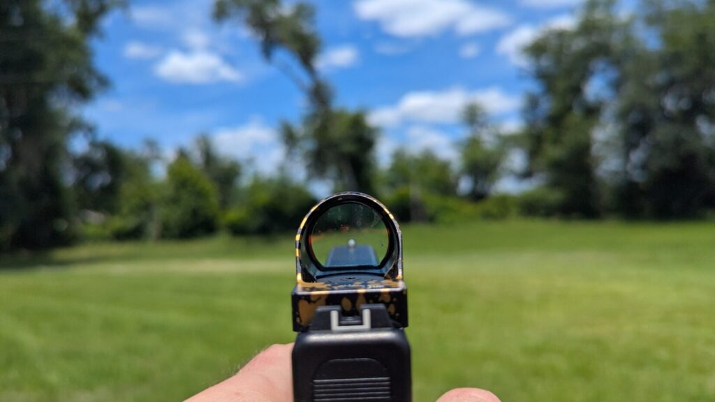 Point-of-view look through a gold-splattered red dot optic with a large, round lens window clearly displaying a prominent bright red dot against an outdoor backdrop.