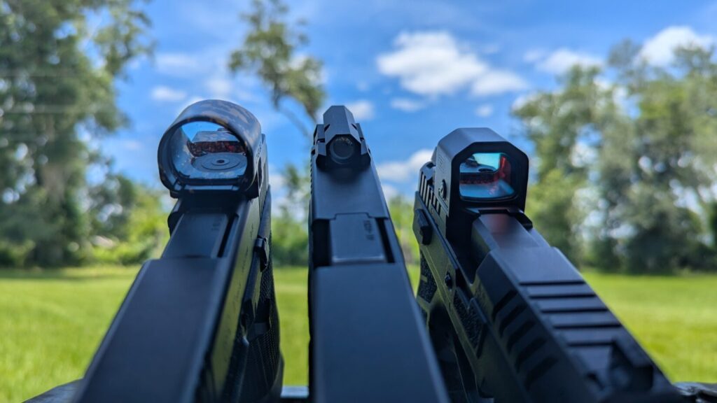 Rear view looking down three handgun slides side-by-side, directly contrasting a large round lens window, a tiny circular tube sight, and a medium square lens window.