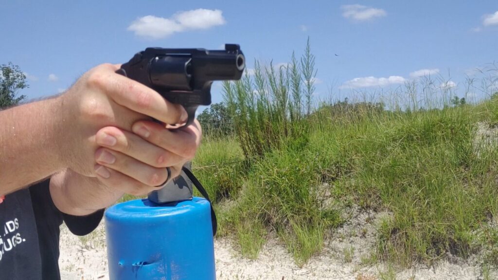 A close-up of a shooter's hands gripping a black snub-nosed revolver at an outdoor range, with a blue plastic barrel visible below.