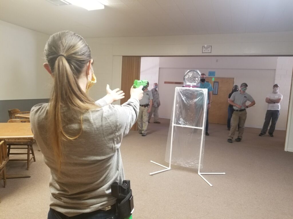 Image of a woman with a ponytail, facing away from the camera in the foreground, turning a palm up as if exasperated. In her other hand she holds a green pistol replica with a laser attachment. The laser is shining through a cellophane replica of a man. Even through her pistol and laser are aimed at the head of the see-through threat target, it is not pointed up far enough, while she stands, to clear the tall bystander behind. A bright green dot is on the wall behind and just off to the side from the tallest bystander. A crowd of other bystanders is behind the cellophane threat target.