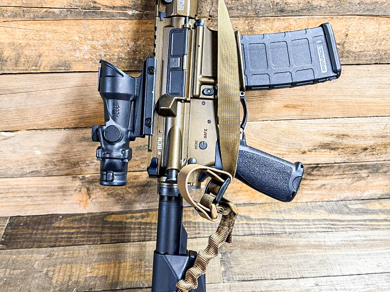 A bronze AR-15 with a black ACOG mounted to it, leaning against a wood wall.