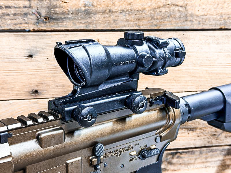 A front/side view of a black ACOG optic mounted on a bronze AR-15.