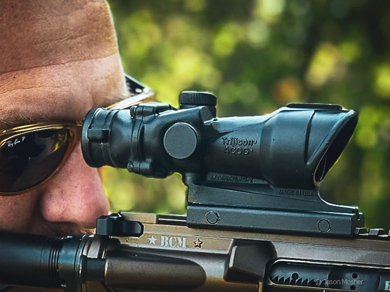 A person in sunglasses shooting a rifle, but the image is zoomed in on the black optic mounted to the rifle.
