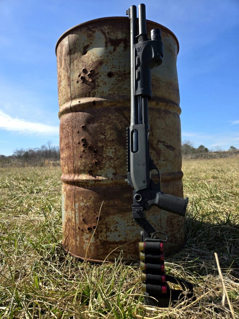 A shotgun next to a 55 gallon drum in a field with a blue sky