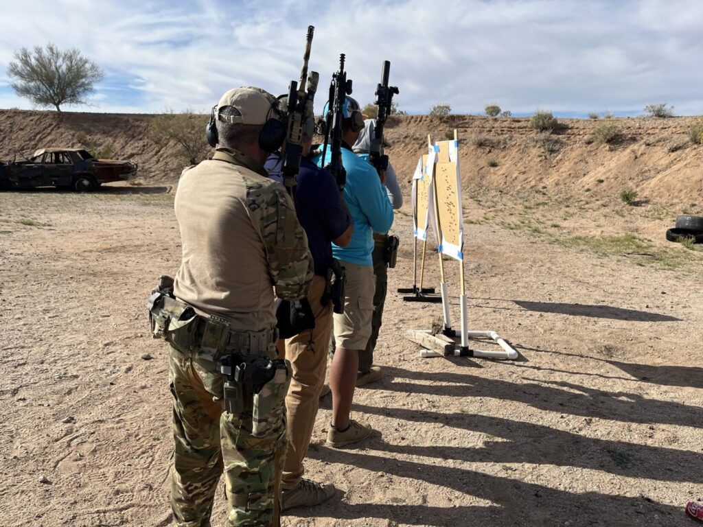 Image of trainees stacked one behind the other, getting ready to go through a simulated door on a firing range. The rescuers behind the one ifn front all have the muzzles of their rifles pointed up for safety.