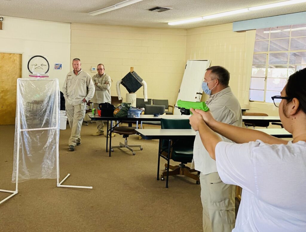 Image of a student pointing a replica pistol with a lser at a cellophane target. Instructors in tan shirts observe.