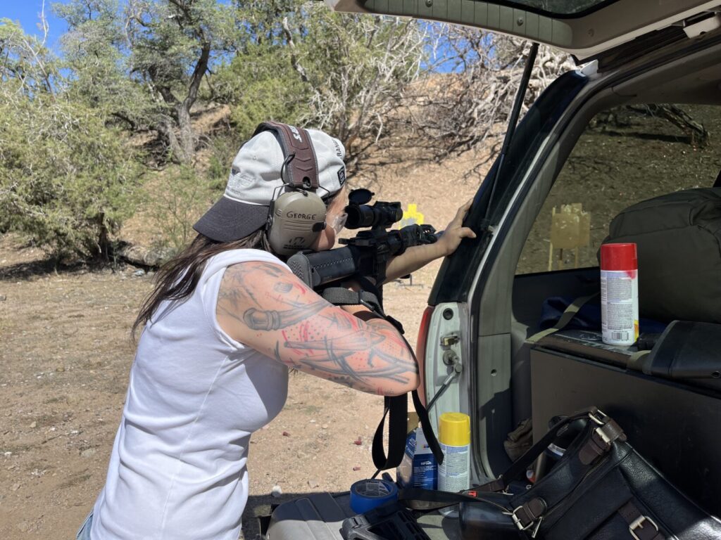 Image of a woman with her hat on backwards shooting a scoped rifle. Downrange is a steep hillside. She's resting the front of the AR-15 on her left forearm. He left hand is braced on the back corner of an SUV.
