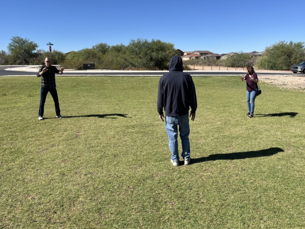 Three people in a park. A potential assailant in a hoodie sweatshit faces away from teh camera. On the left, and farther away, a man with a beard faces the potential assaialnt with his hands out in front of him. A woman on the right looks over he shoulder to scan for accomplices.