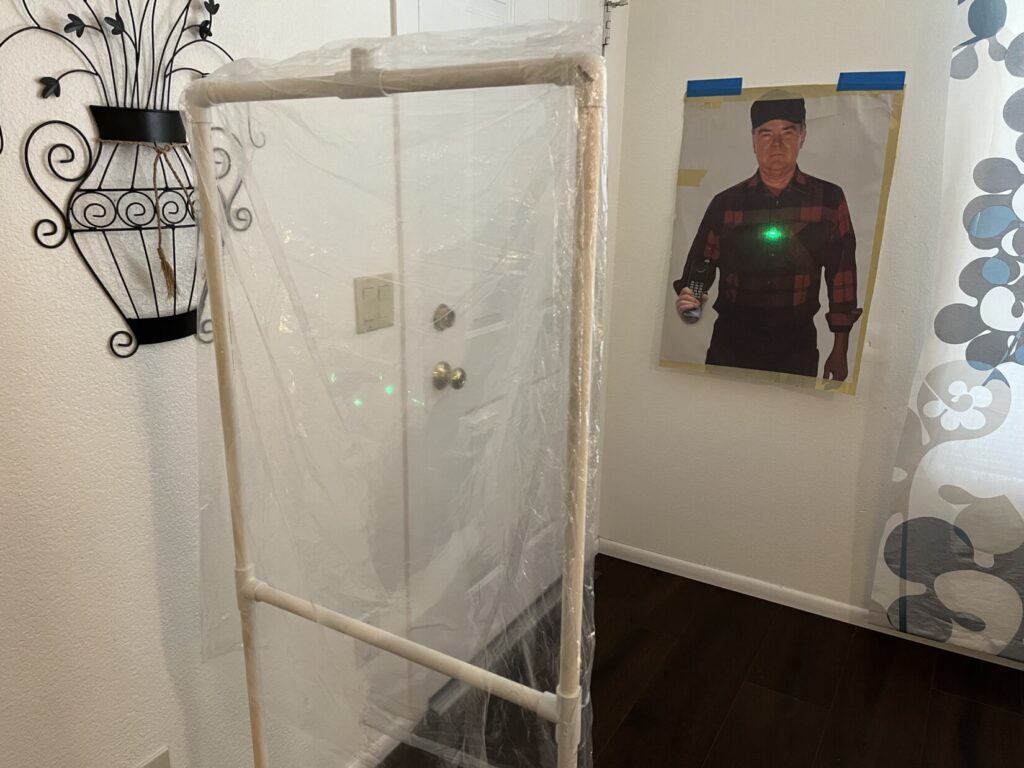 Image of a white PVC pipe frame with a dry-cleaning garment bag over it. The middle of the garment bag glows with refracted green laser light. Behind the target on the wall is a man in a flannel shirt holding a Motorola radio in his right hand. The green laser, having passed through the cellophane, splashes a bright green dot on the chest of the hiker in the flannel shirt.