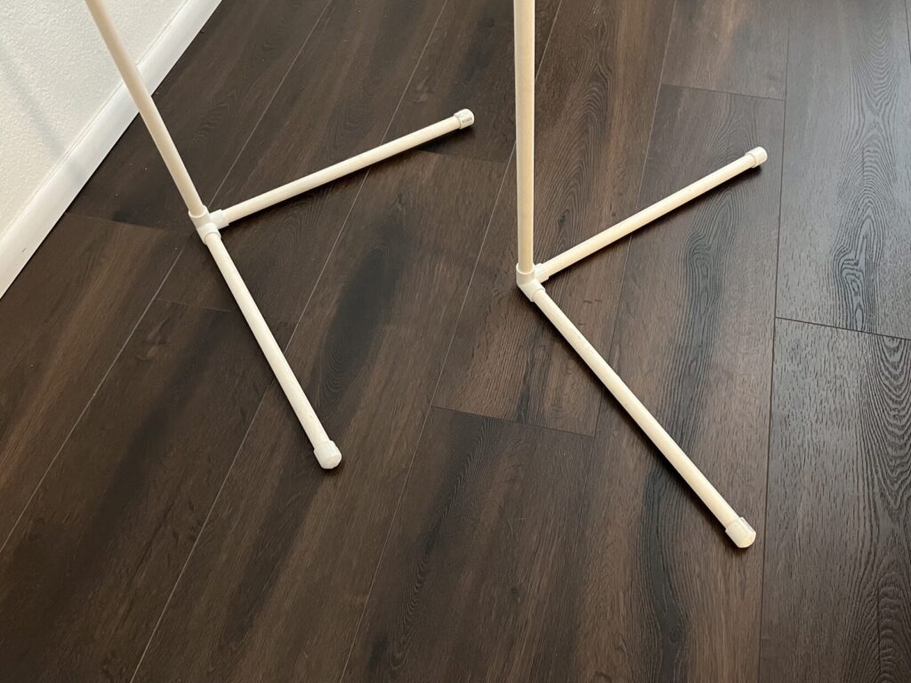 The white PVC pipe legs of the Cellophane Man frame are pictured going down to a brown plank floor. Each leg has two straight sections of PVC pipe, called feet, jutting out of it at a right angle forming a capital L. The two Ls are arranged like chevrons so the frame can be placed next to a wall.