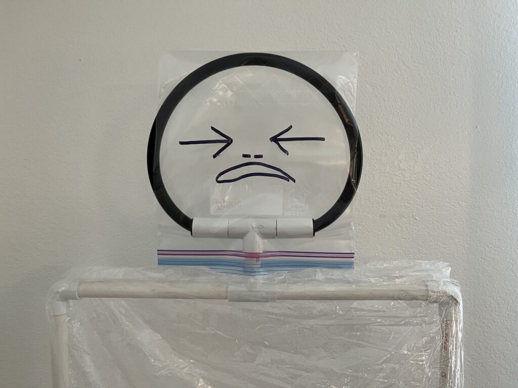 This image shows the cartoon-like face of Cello Man, drawn with a fat magic marker onto an upside down, gallon-sized Ziploc bag. The bag is stretched over a hoop made from a semi-rigid plastic pipe fitting. The head is connected to the white PVC shoulders through a pair of T-shaped fittings.