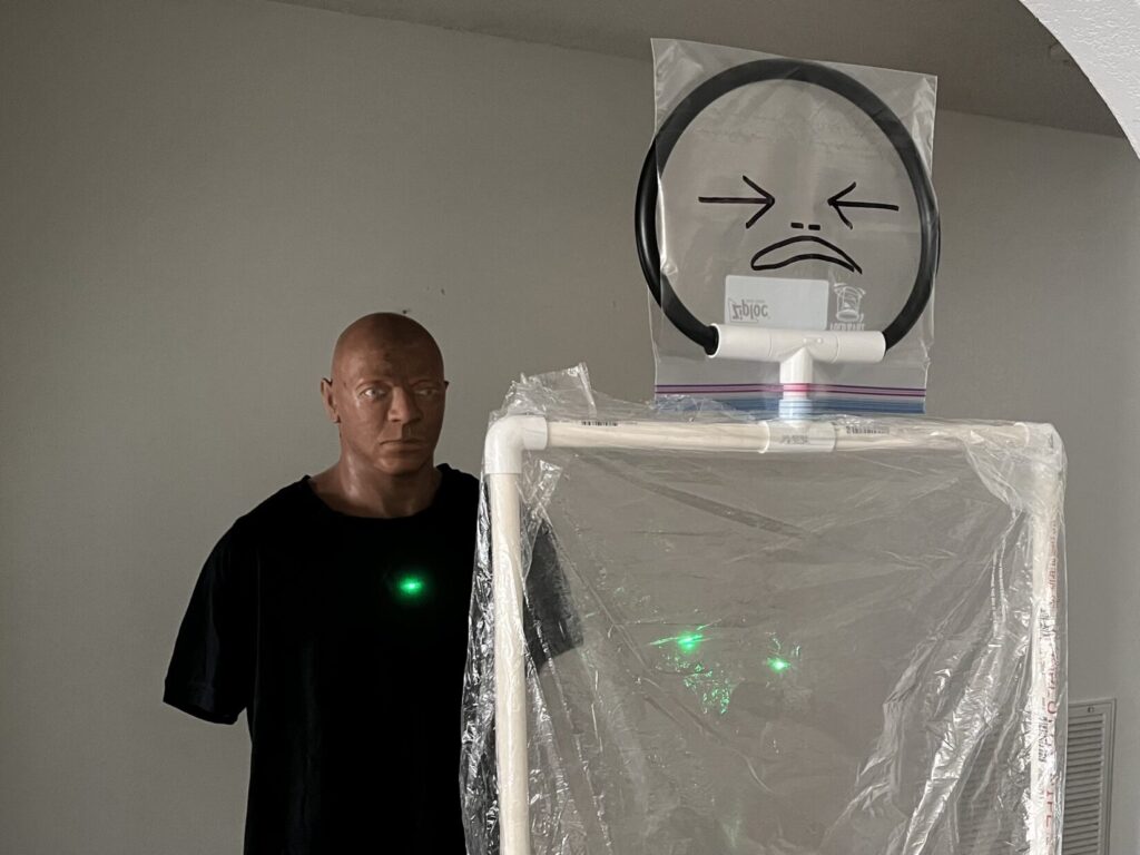 Image of a manikin in a black T-shirt behind a cellophane target. The manikin represents an innocent bystander. The cellophane target represents a killer in the process of murdering innocents. A green laser dot goes through both layers of cellophane and onto the bystander behind.