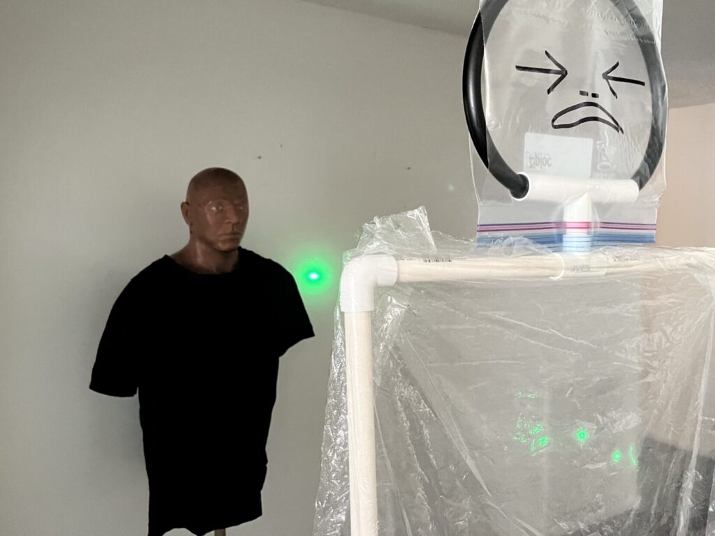 Image of a manikin in a black T-shirt behind a cellophane target. The manikin represents a bystander and the cellophane target represents a lethal threat. A green laser os shooting through the chest of the cellophane target at such and angle that it hits the wall beside the bystander behind.