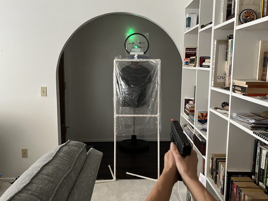 A cellophane target stands in an archway leading from a foyer to a living room. A minikin representing a family member of the defender wears a black T-shirt in the foyer behind the cellophane threat. The hands of the defender are visible in the foreground, holding a Glock model 19 pistol. The pistol has an orange plastic barrel insert sticking out of the muzzle. A laser attached to the pistol shoots a green dot onto the head of the manikin, and through it onto the wall above the innocent bystander's head.