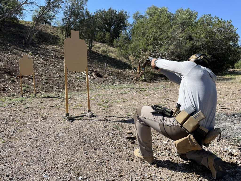 A man in a grey long sleeved shirt kneels and aims at the head of the closest of two silhouette targets. The angle of his pistol is high enough that if he shot, rounds might fly over the hill behind the targets.