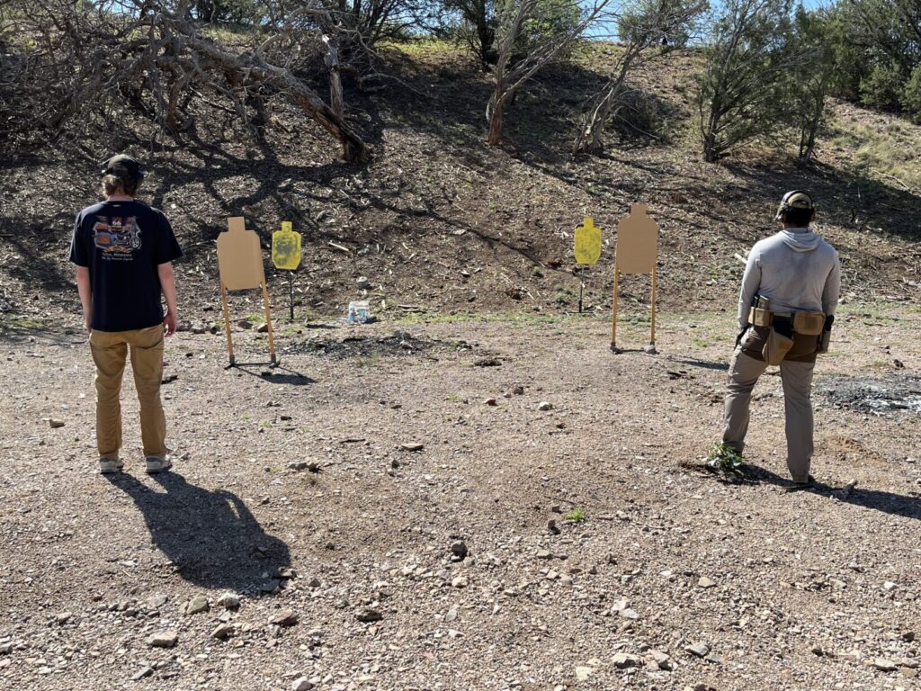Two shooters stand in front of one pair of targets each. The closest targets are cardboard. The farther targets are yellow-painted steel.