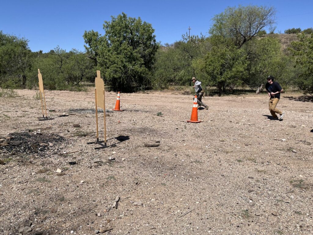 Two training partners run forward and to their right (to the left from our point of view). Two cardboard targets are visible behind them, but the steel targets behind those are off-camera to the left. Orange traffic cones tell the students where to stop so one doesn't get ahead of the other.