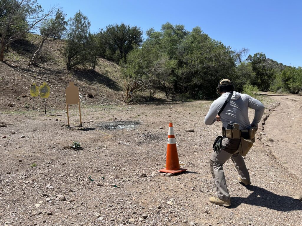 A man starts in front of a line of targets. The closest is cardboard. There is a yellow-painted steel target behind that 10 meters away. Another steel target is about 3 meters behind that. The shooter is reaching for his pistol as he moves to get a clear line of fire on the designated target.
