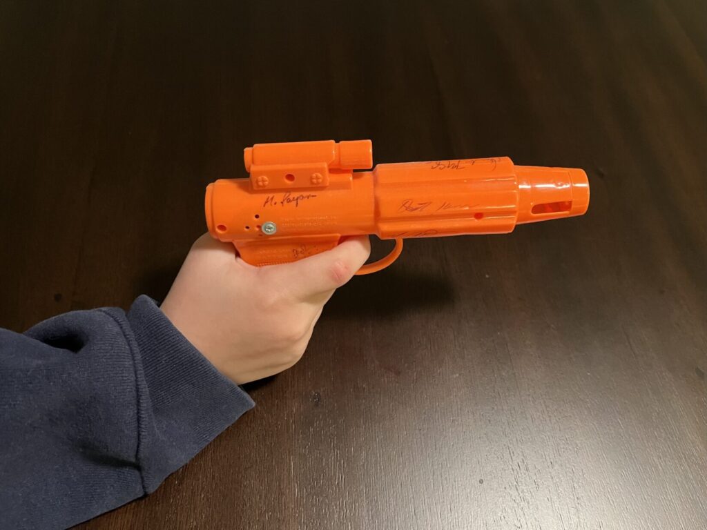 Image of an orange plastic toy shaped like a science fiction laser blaster. A hand holds the toy with the index finger on the trigger.