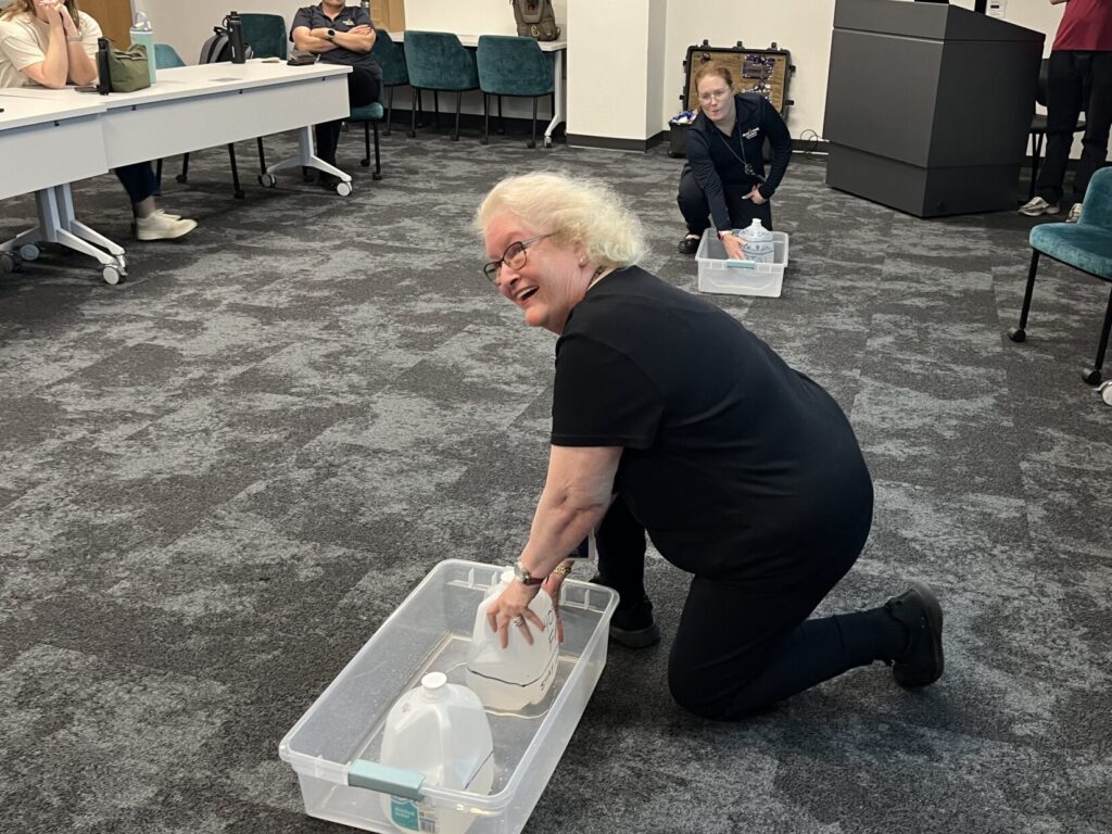 A woman holds direct pressure on the side of a jug set up next to a half-gallon jug in an open plastic tub on the carpeted floor inside a classroom. In the background, another woman holds pressure on the side of a one-gallon jug that started eight-tenths full. That jug is also in a wide, open-topped plastic bin.