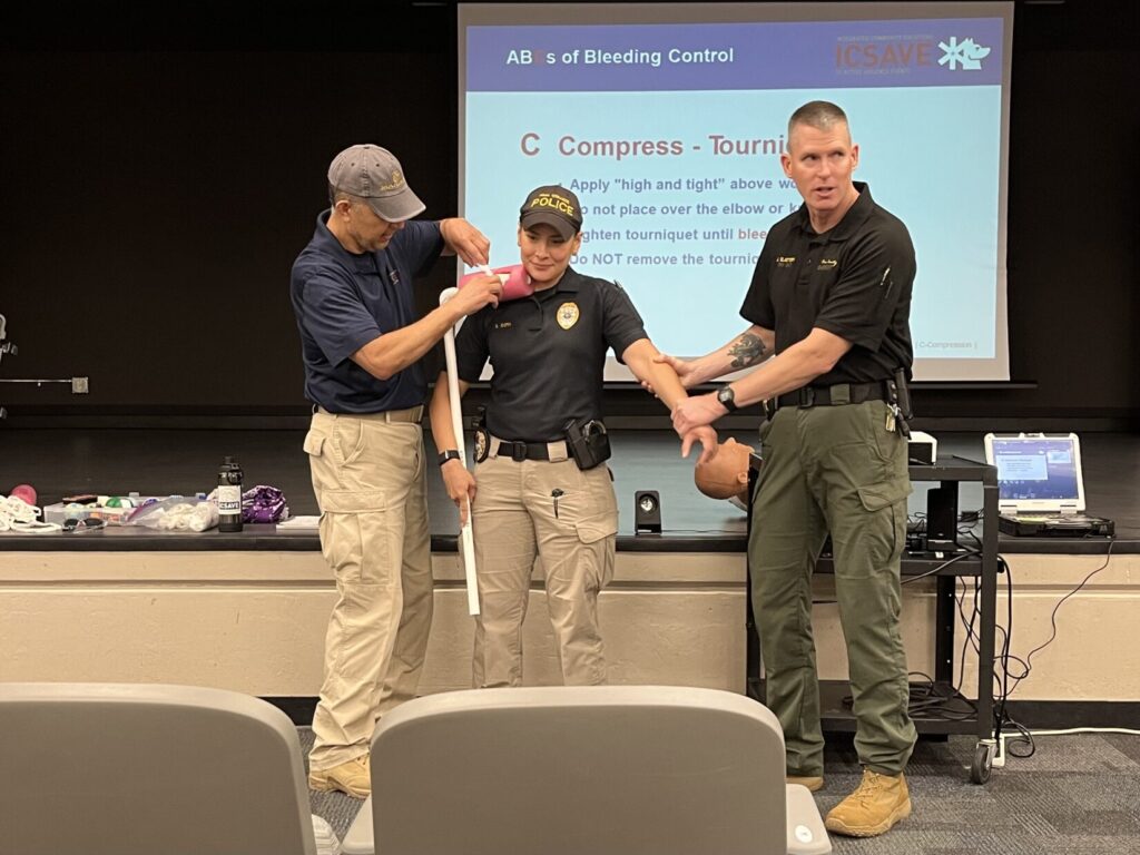 A Bleeding Control instructor demonstrates how to pack gauze into an open wound under the collar bone. A role player pays a patient, standing up so she can be seen by an audience. Another instructor holds her opposite wrist to demonstrate that packing wounds is painful and the patient may struggle.