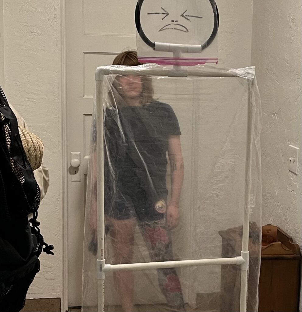 Image of a short woman in a black T-shirt, holding a pistol trainer with one hand, by her side, pointed att he ground. Her image is mostly obscured by the translucent dry-cleaning bag of the Cellophane Man training aid.