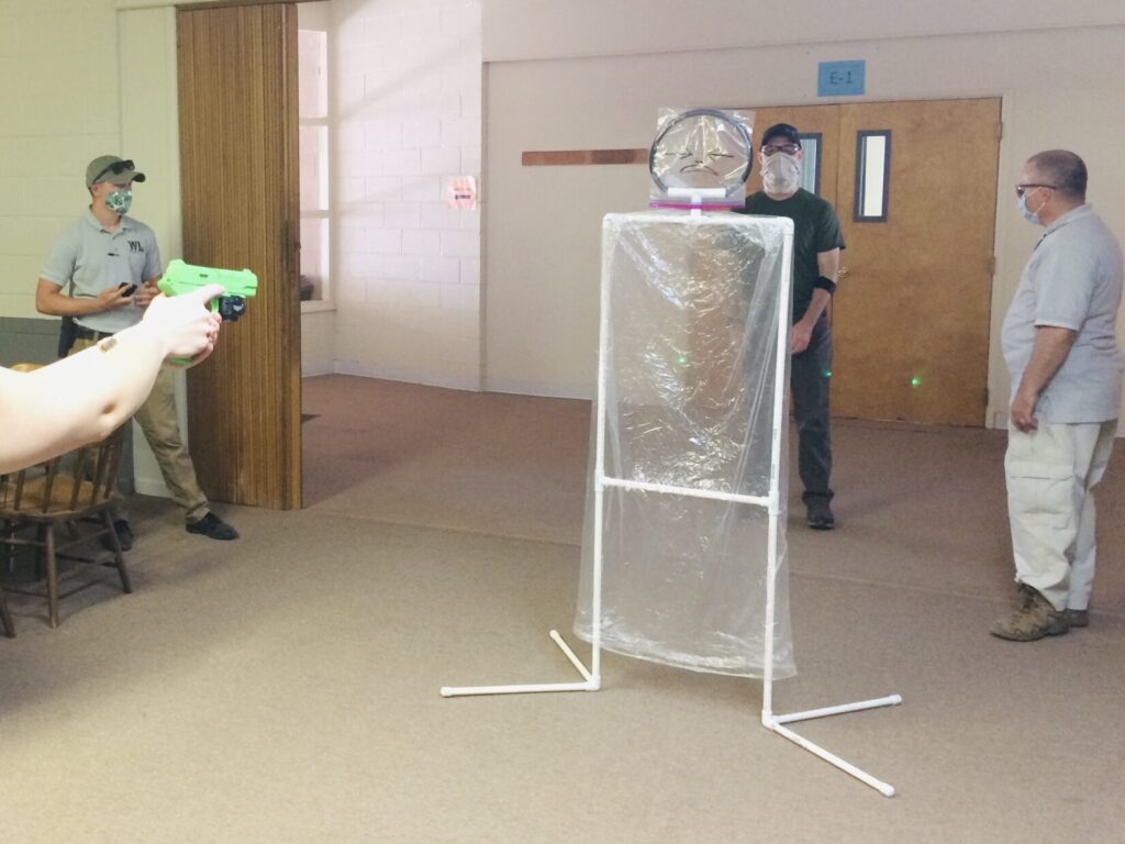 A student aims a green laser though a cellophane target representing a threat. The laser is also glancing off the edge of the leg of a bystander role player behind and continues on to the bottom of a door behind that.