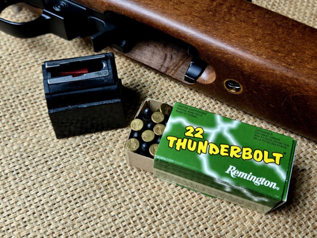 A box of rimfire ammunition, box magazine, and rifle against a burlap background