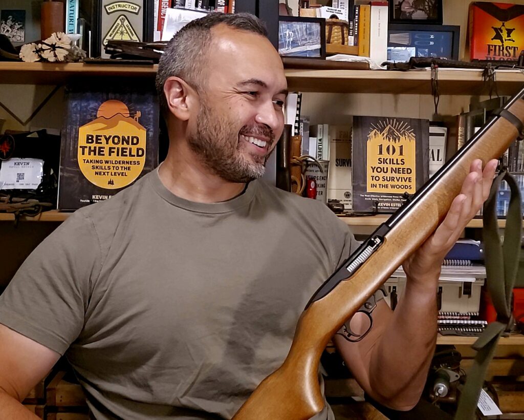 A man in a green shirt sitting in front of a book case looking at a rifle.
