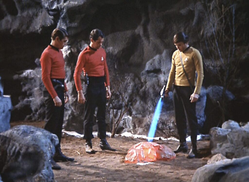 Image from the original Star Trek TV series of Chekov in a yellow shirt using a phaser to warm a rock on a cold planet. Two expendable red-shirted security personnel stand nearby.