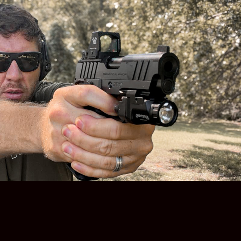 A person with sunglasses, shooting a 1911 handgun outdoors, with trees in the background.
