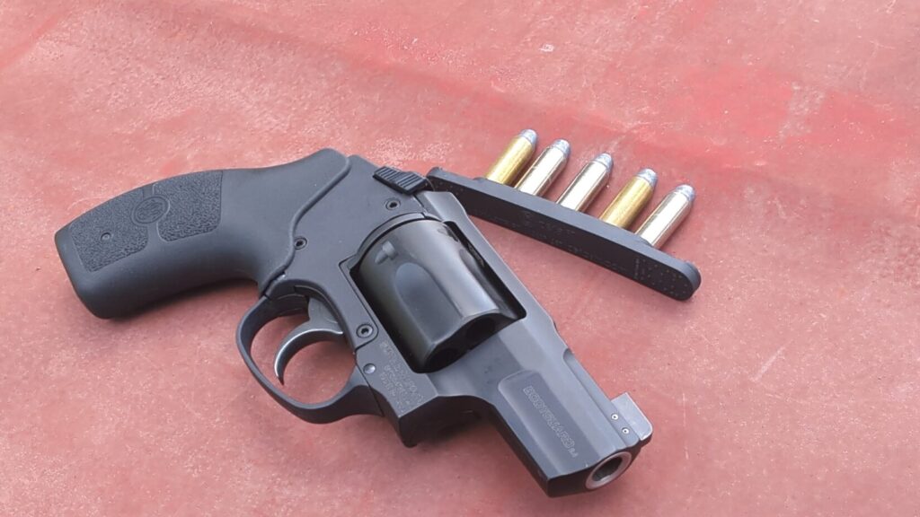 A black compact revolver labeled "BODYGUARD 2.0" rests on a textured red surface next to a black speed strip holding five cartridges.