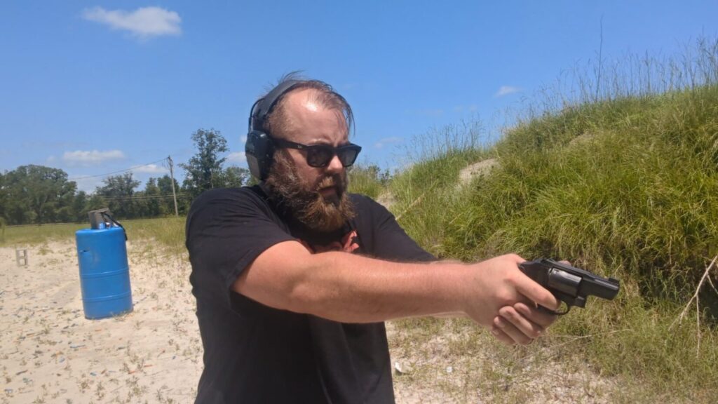 A side view of a bearded man wearing sunglasses and ear protection while firing a small black revolver at a sandy outdoor range.