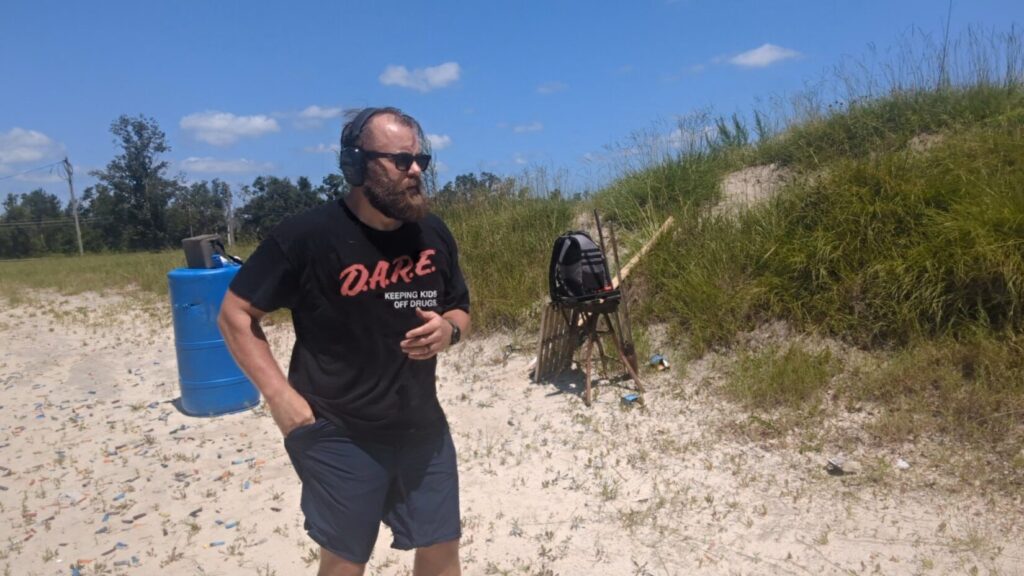 A bearded man wearing a black "D.A.R.E." T-shirt and ear protection walking across a sandy outdoor shooting range.
