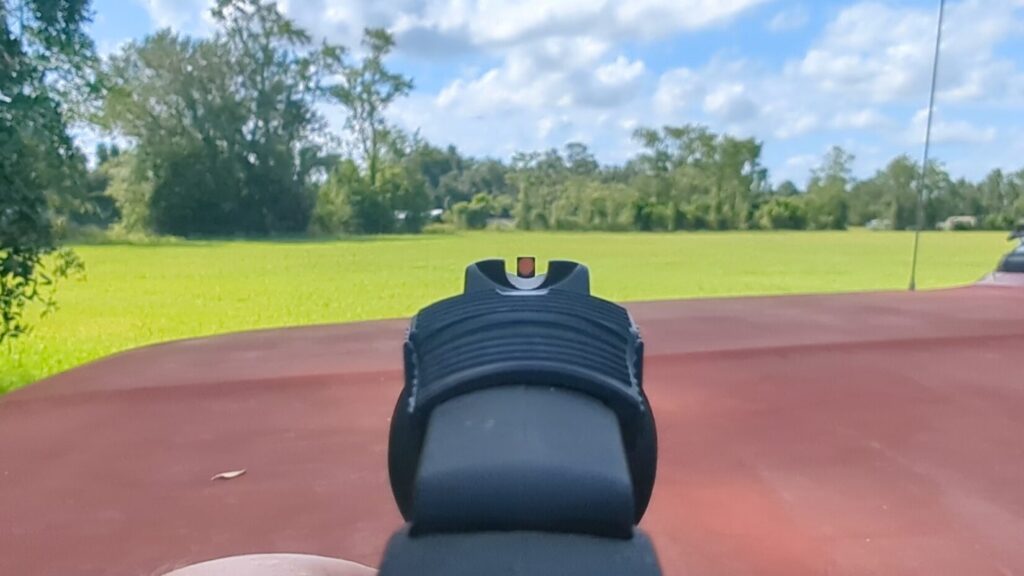 A first-person view looking down the ribbed top strap and bright orange front sight of a revolver, aimed toward an open green field.