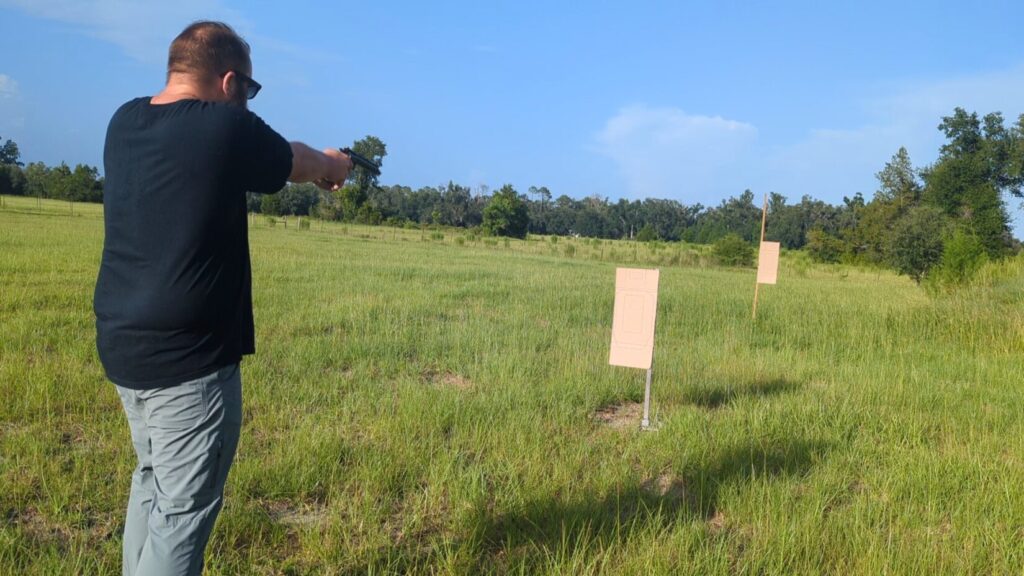 A view from behind of a man in a black shirt and gray pants aiming a handgun across a large, open green field with cardboard targets.