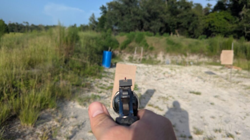 A first-person point-of-view perspective looking down the barrel and iron sights of a revolver aimed at a cardboard target on a sunny, sandy range.