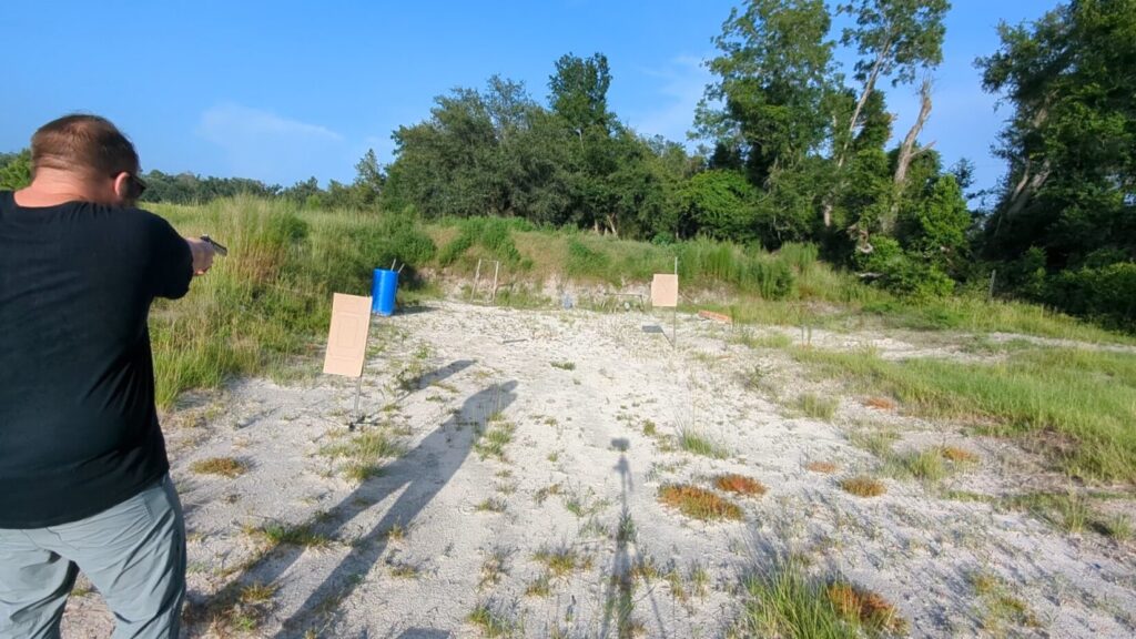 A man aiming a black revolver at an outdoor sand-and-gravel shooting range with trees in the background.