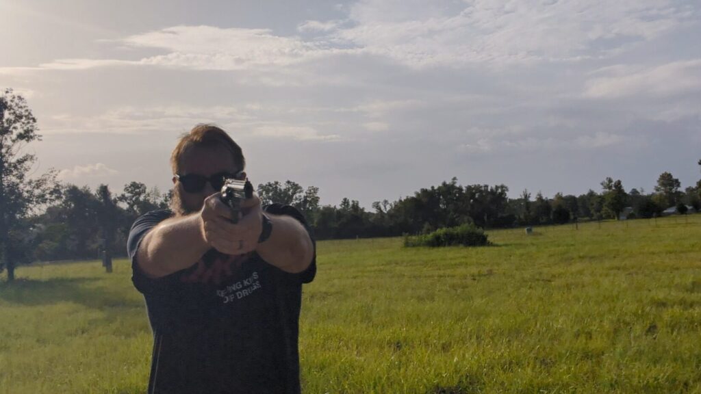 A front-facing view of a man wearing sunglasses aiming a revolver directly toward the camera against a field and cloudy sky.
