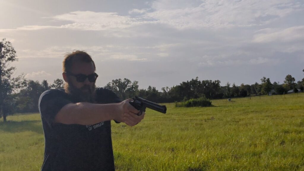 A side-profile view of a bearded man wearing sunglasses, holding a long-barreled black revolver with both hands in a grassy field.