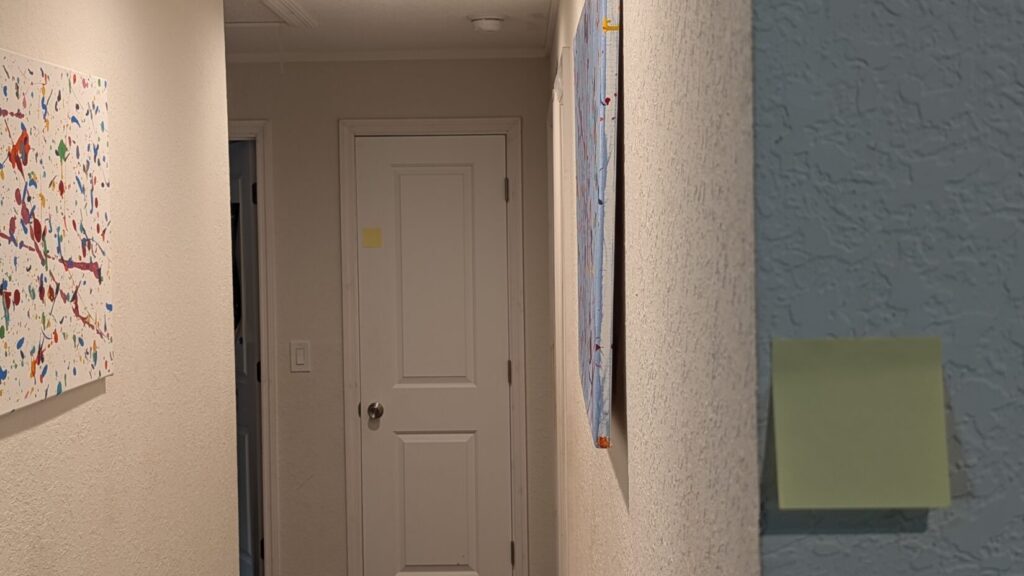 A residential hallway featuring a white door, colorful splatter-paint canvases on the walls, and square sticky notes attached to the wall and door frame.