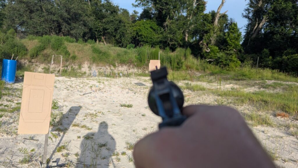 A first-person view focusing on the rear sight alignment of a revolver, pointing toward a cardboard target on an outdoor training range.