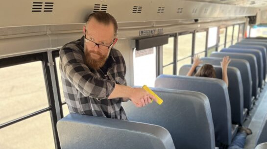 A man stands in the row of a bus with his back to a window. He's pointing a yellow plastic pistol replica at a person pretending to be an assailant, who is prone in the aisle. A woman a few rows ahead of them keeps her head down with her wrists on the top of the seat in front of her, so her hands are visible. The man with the yellow pistol is looking away from them, scanning for potential accomplices of the man down in the aisle.