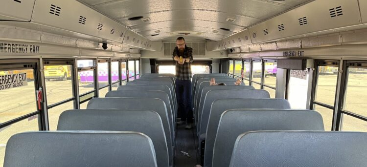 A man stands in the aisle, all the way to the rear of a bus. He's holding a yellow plastic pistol replica, pointing it at a man in a hoodie who is prone in the aisle. A knife is on the floor between them. A woman on the right has her face down and her wrists propped up on the seat in front of her.