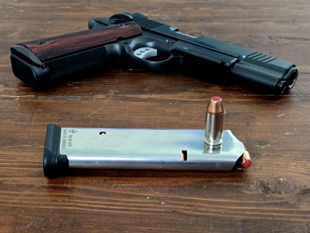 A 1911 pistol on a table with a loaded magazine in front with one round of ammo on top.