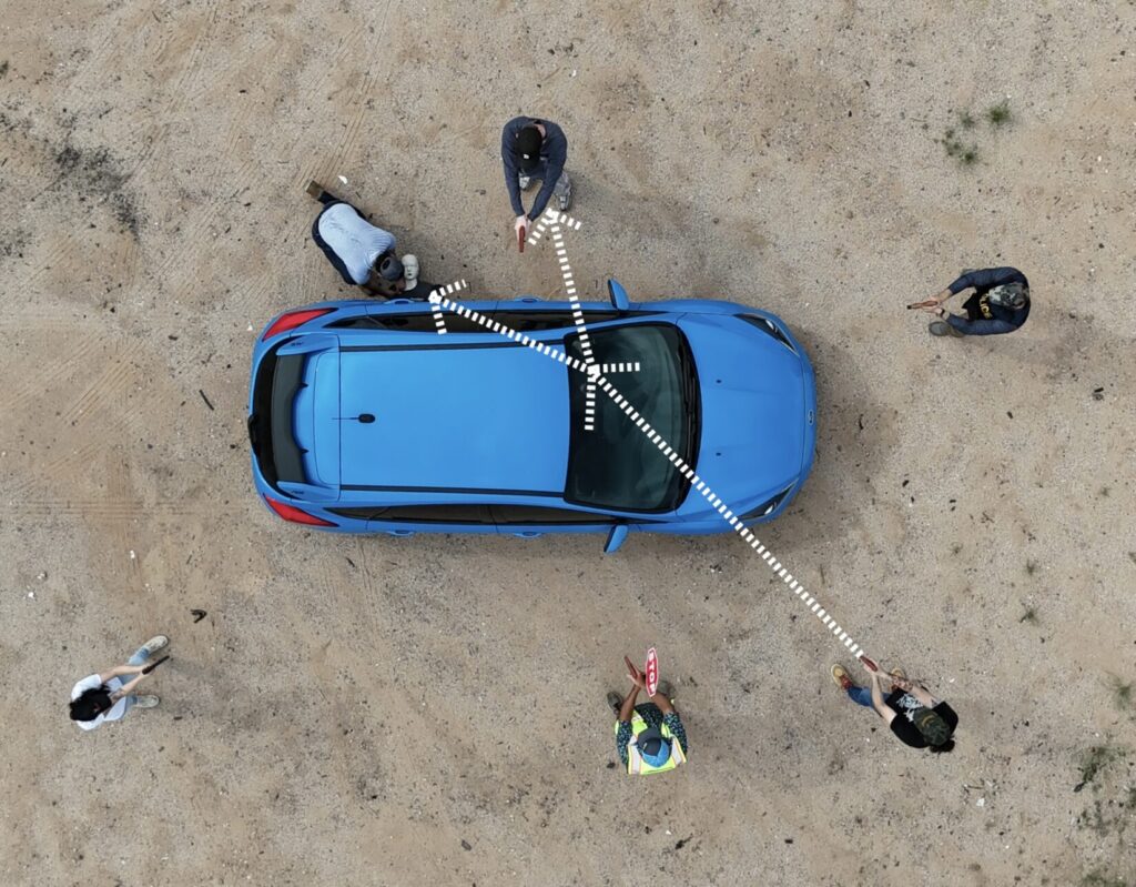 Image of a blue car from above. Role players point plastic pistol replicas at the car. Arrows indicate the possible paths a bullet might take, hitting teammates on the other side.