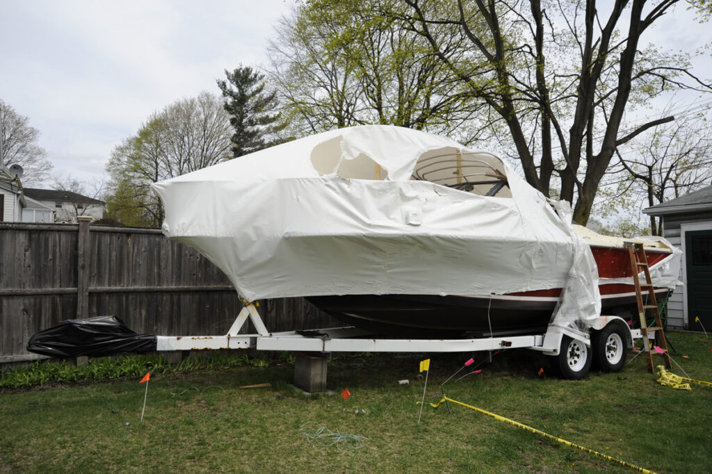 View of a bullet-riddled red boat with a torn white cover on a trailer in a back yard.