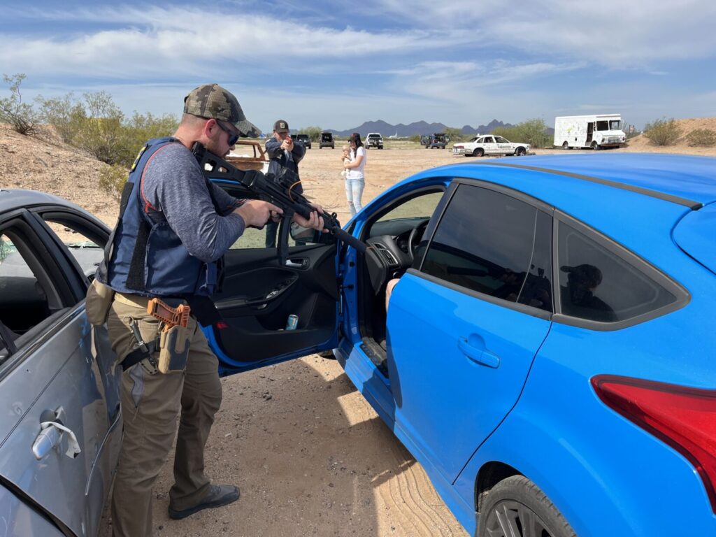 A role player in Police raid gear re-enacts how special agent Sweetow was standing next to the open door of the sedan while his partner stood in front of the sedan, pointing through the windshield. A concerned woman holds an infant CPR manikin representing her baby.