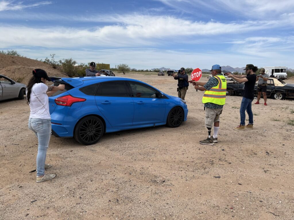 Several persons are standing around a small blue sedan, pointing plastic replica pistols at it, and each other.