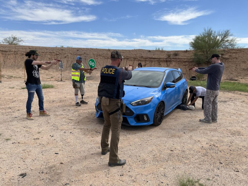 Image of several people, including a school resource officer and a crossing guard, all pointing guns at a car while another rescuer kneels beside manikin representing a youth who had been run over, by the back left tire.